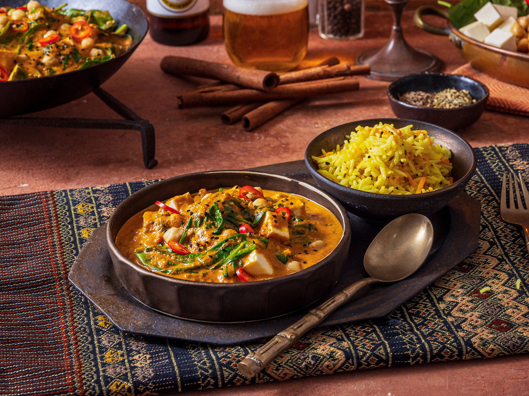 Two bowls of curry and rice on a patterned tablecloth with cinnamon sticks and a glass of beer in the background.
