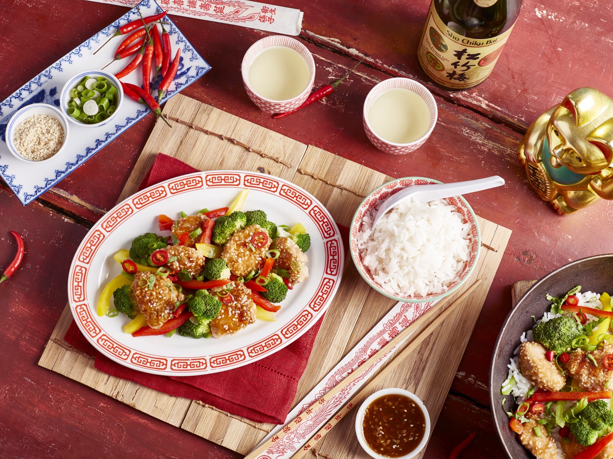 Chinese dish with vegetables and rice on a red tablecloth with chopsticks and sauces.