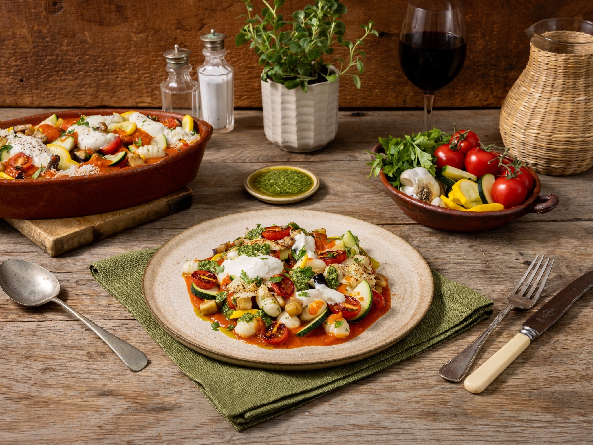 Dish of pasta with vegetables on a wooden table with a glass of red wine and potted plants.