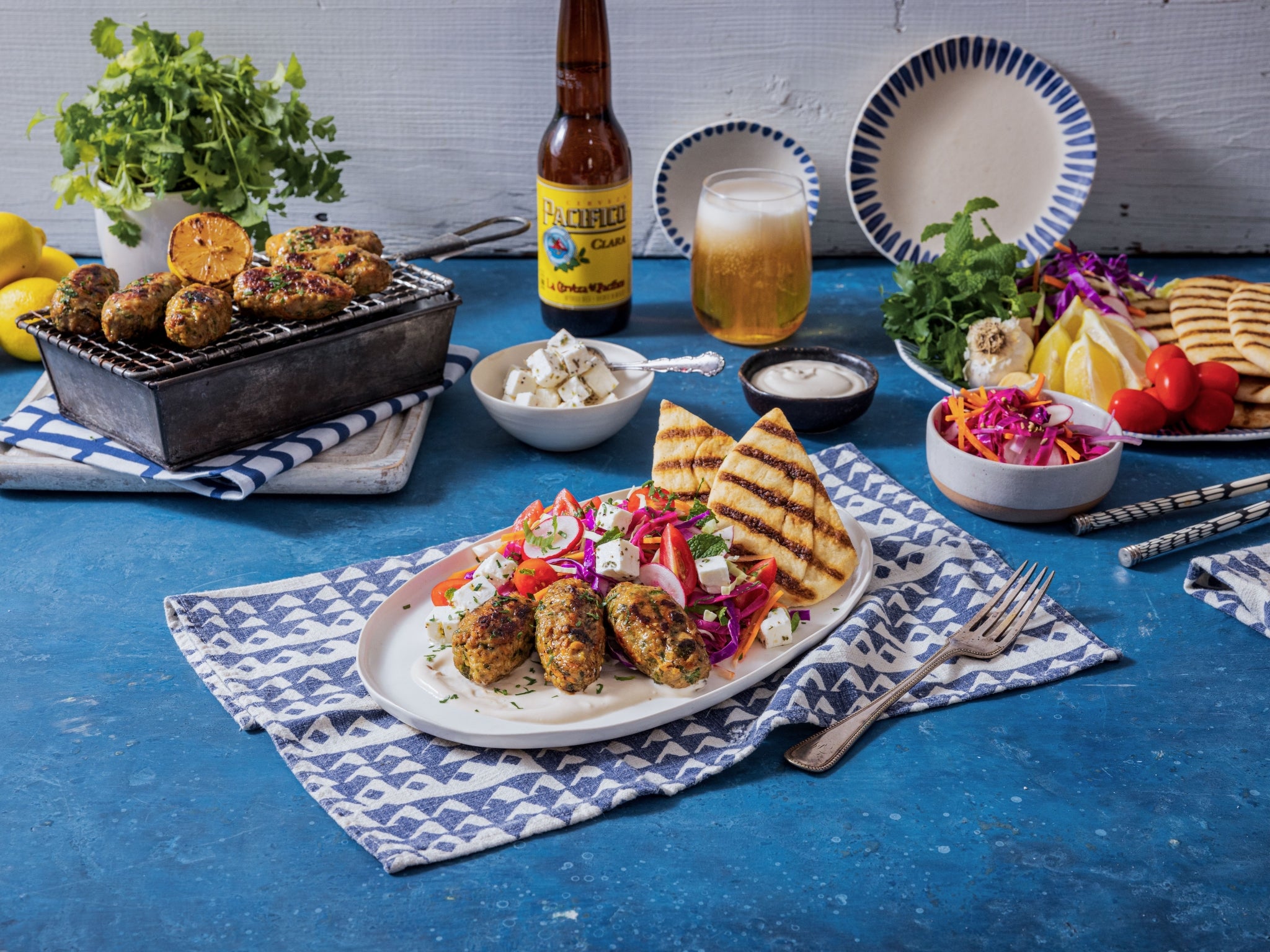 Table setting with grilled food, beer, and side dishes on a blue surface.