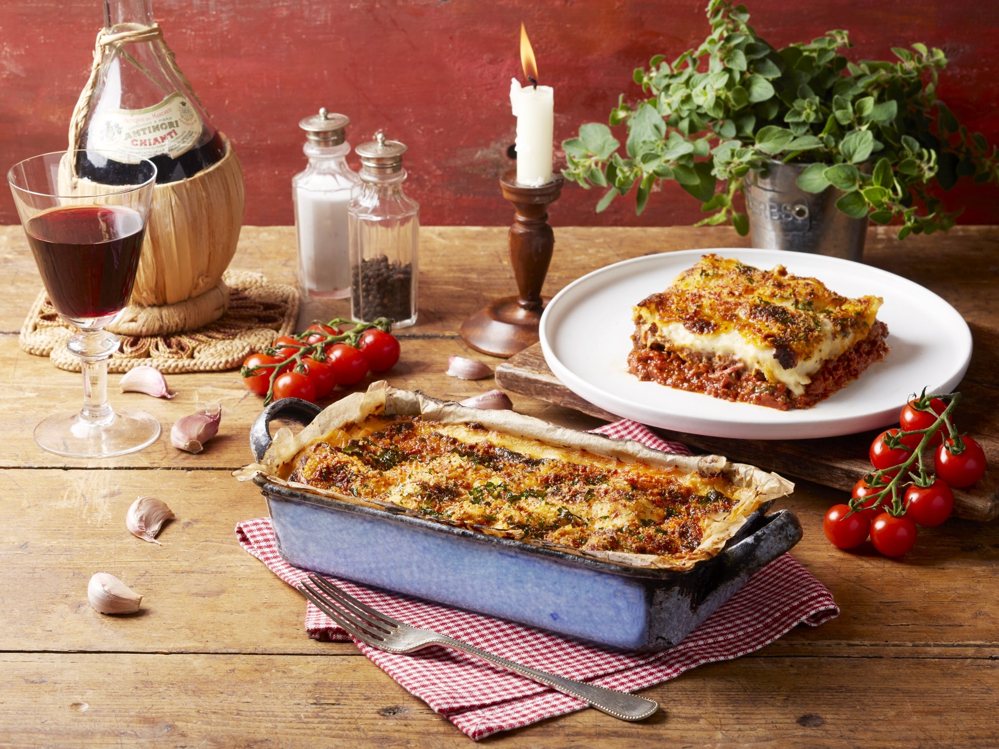 Two dishes of lasagna on a wooden table with wine and herbs.