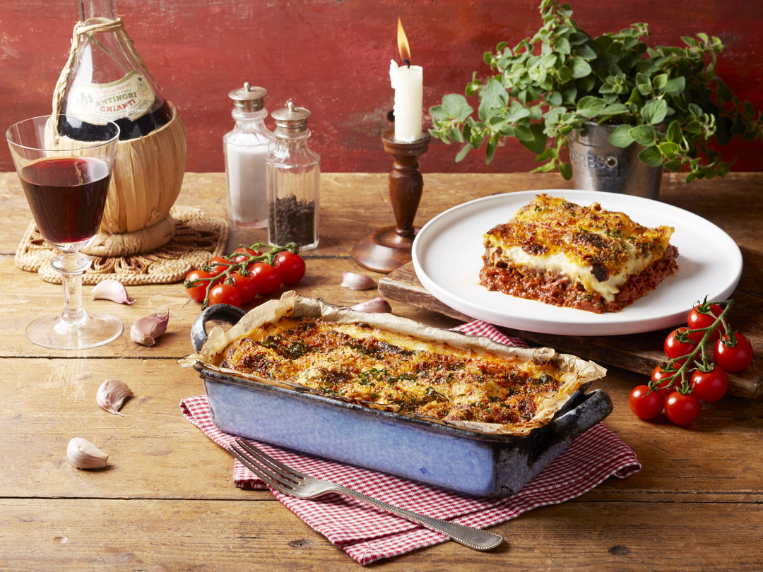 Two dishes of lasagna on a wooden table with wine and herbs.