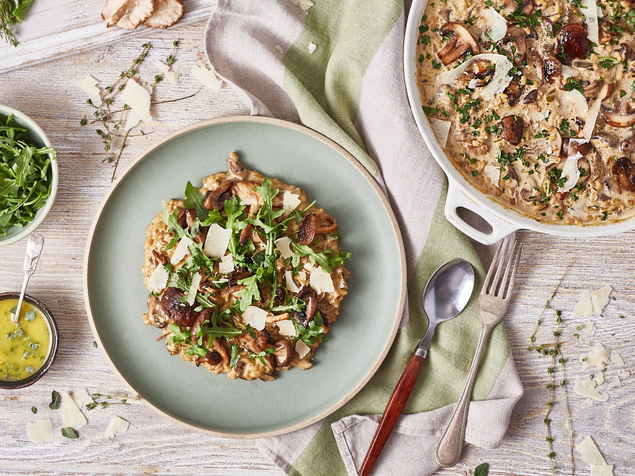 Plated dish of risotto with arugula and parmesan on a wooden table with a pot of risotto and a bowl of greens.