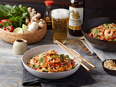 Dish of noodles with shrimp and vegetables on a wooden table with a glass of beer and bottles.