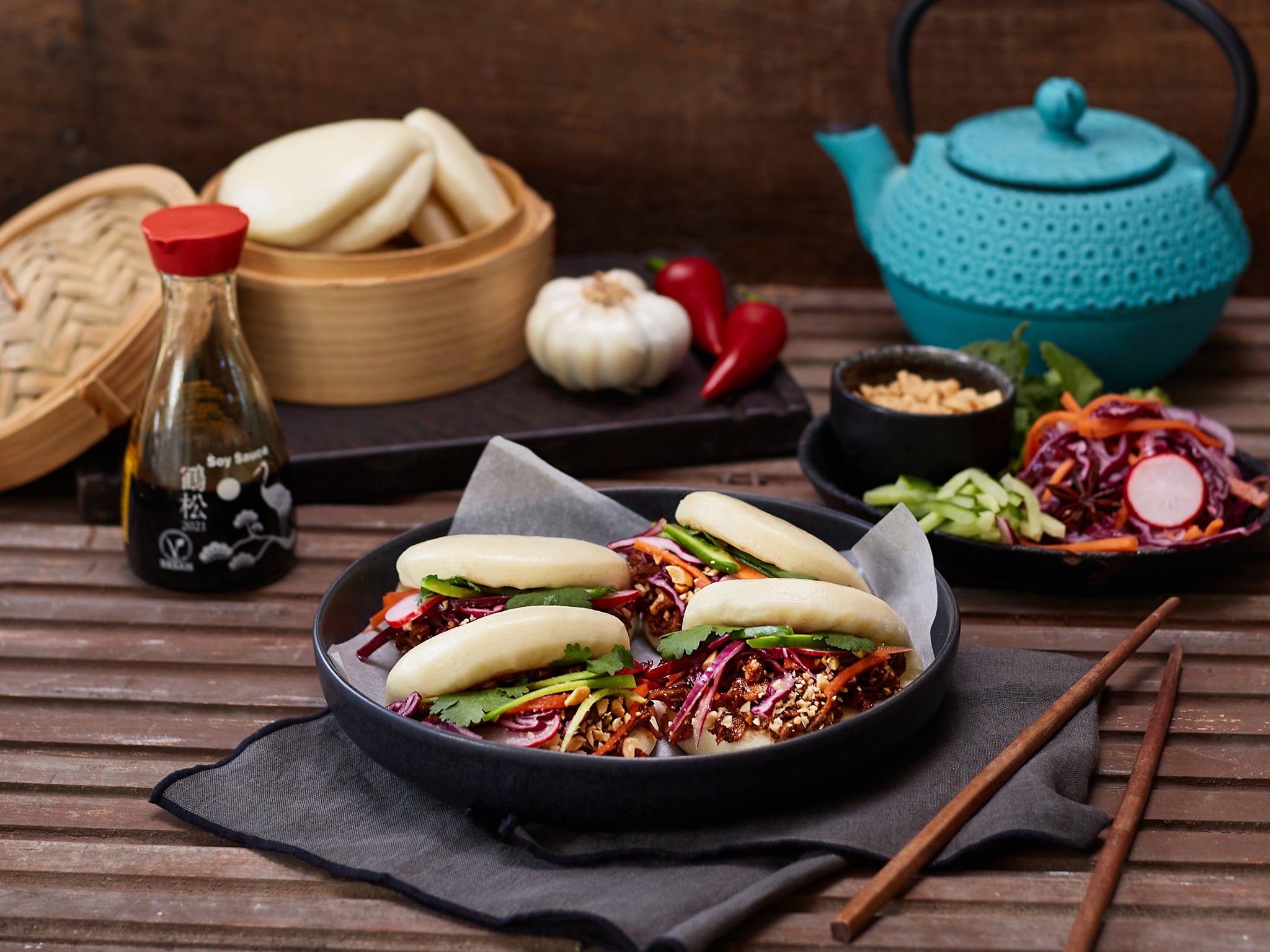 Dish of baozi with vegetables on a wooden table with a teapot and condiments.