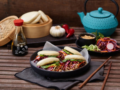 Dish of baozi with vegetables on a wooden table with a teapot and condiments.