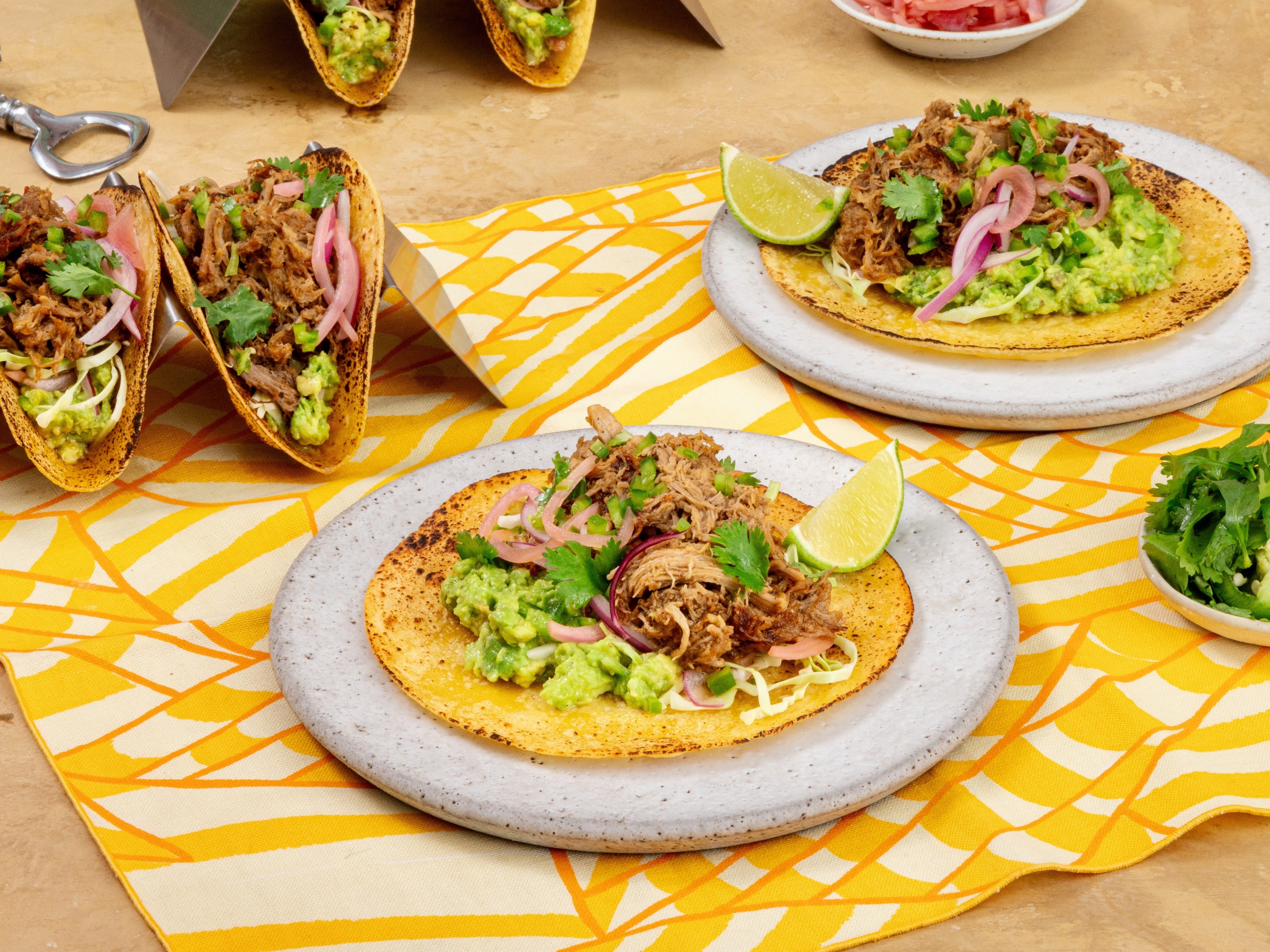 Tacos with meat, guacamole, and lime on a yellow and white patterned tablecloth.