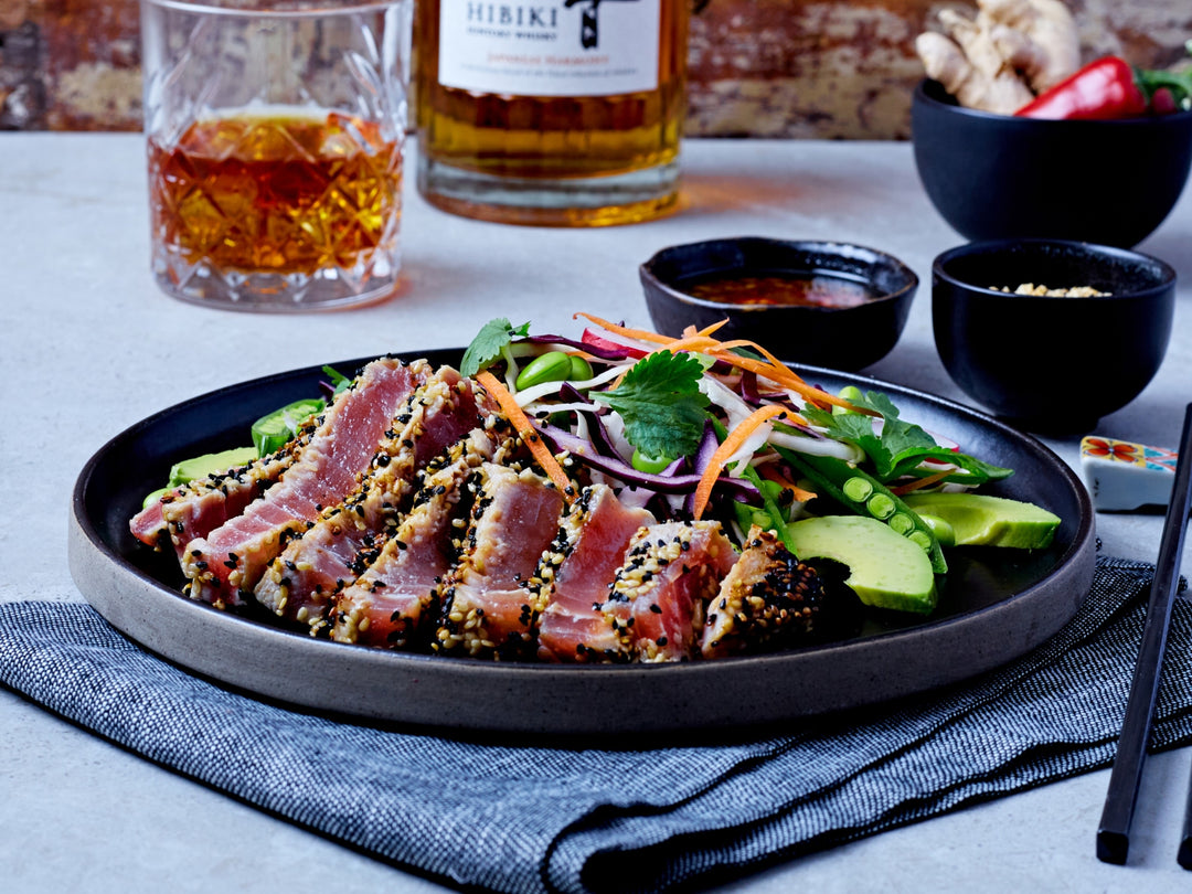 Sliced ahi tuna with avocado and vegetables on a plate with drinks and garnishes in the background.