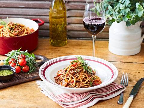 Spaghetti dish on a wooden table with a glass of red wine, tomatoes, and herbs.