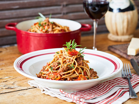 Bowl of spaghetti with a red pot in the background on a wooden table.
