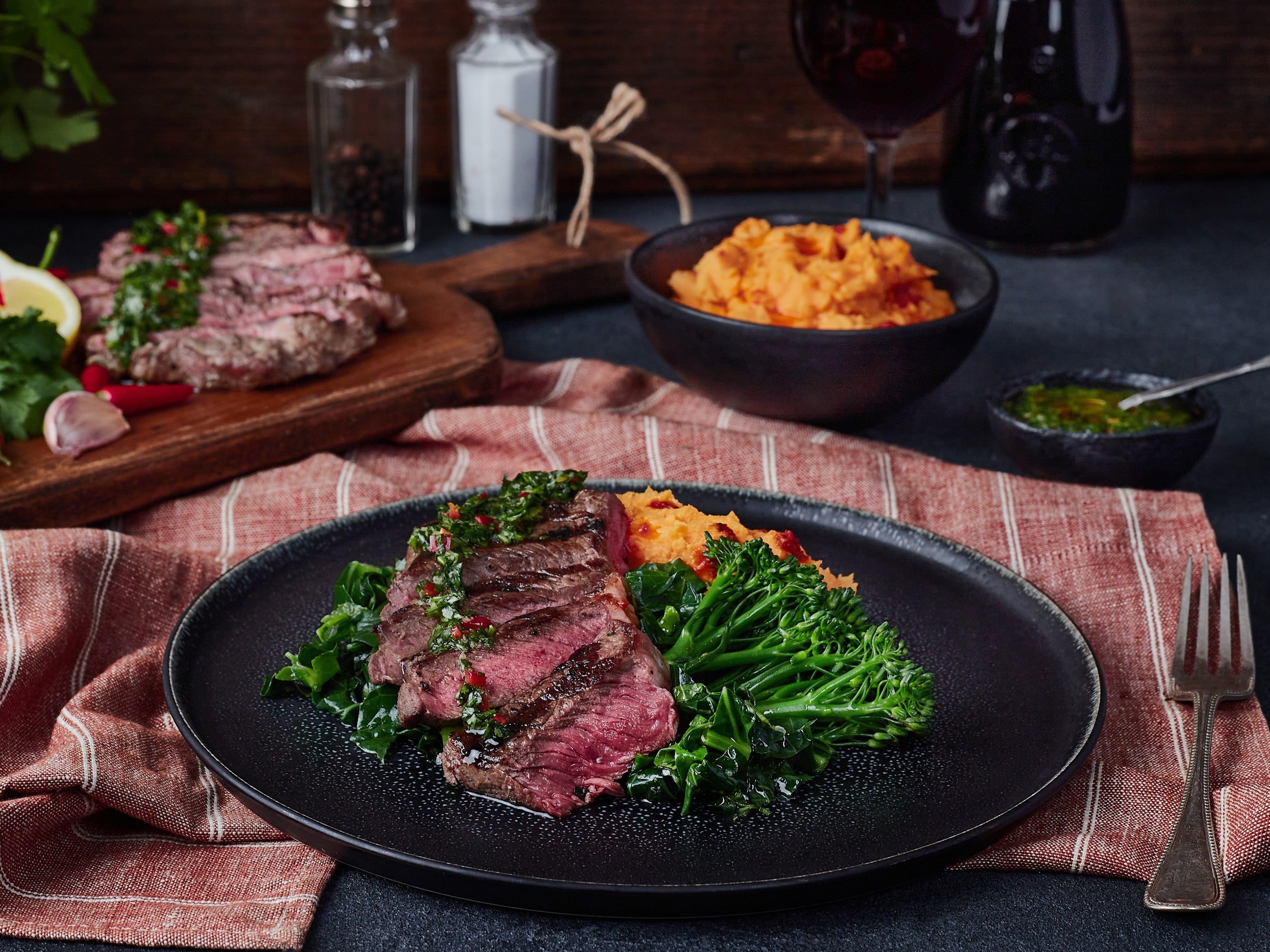 Steak dish with vegetables on a black plate, served with mashed potatoes and a glass of red wine.