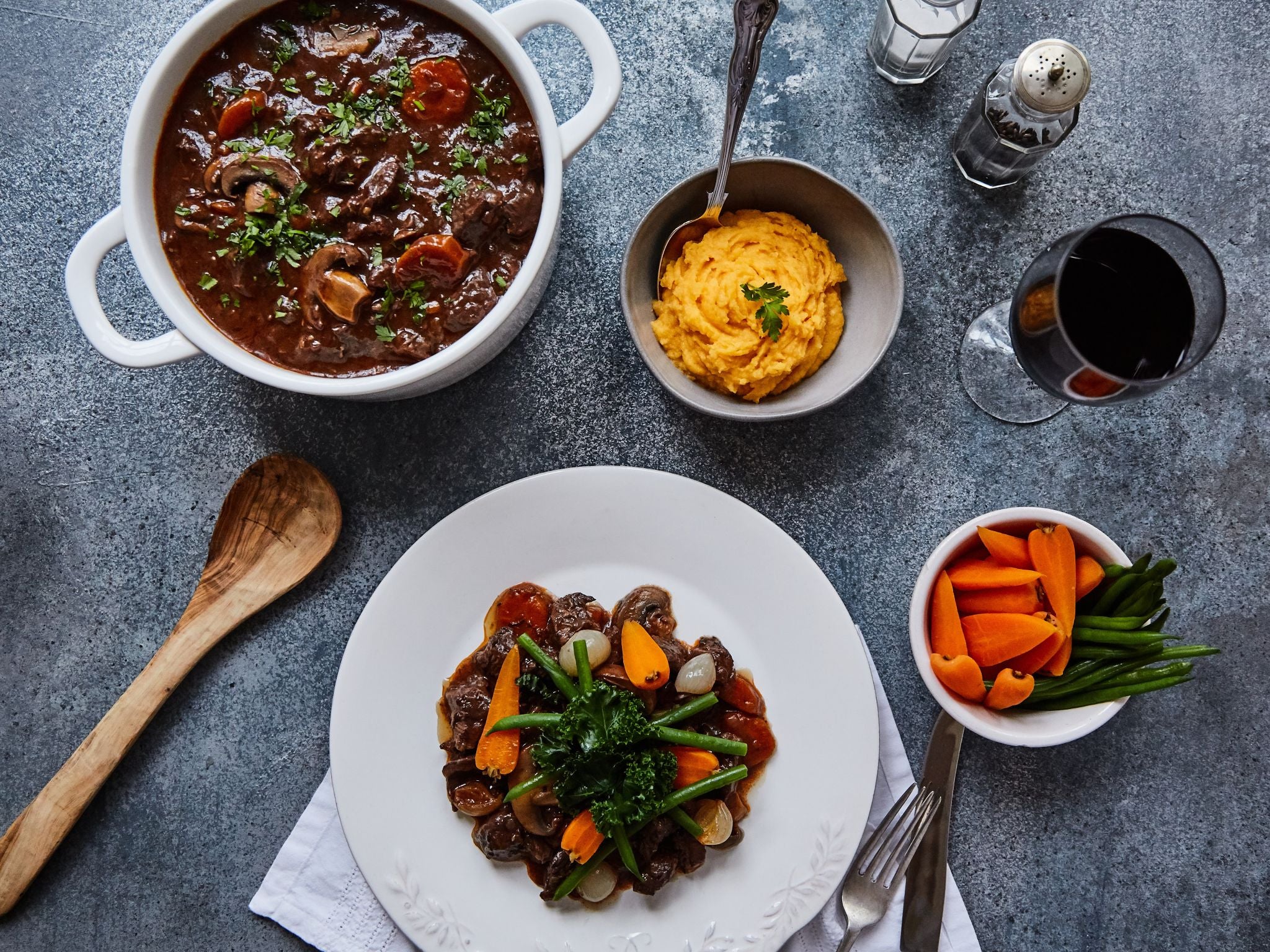 Dinner table with a bowl of stew, a plate of salad, and side dishes on a gray surface.