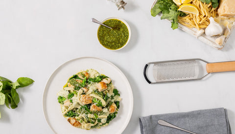 Pasta dish with greens on a white plate, surrounded by ingredients and utensils on a light surface.